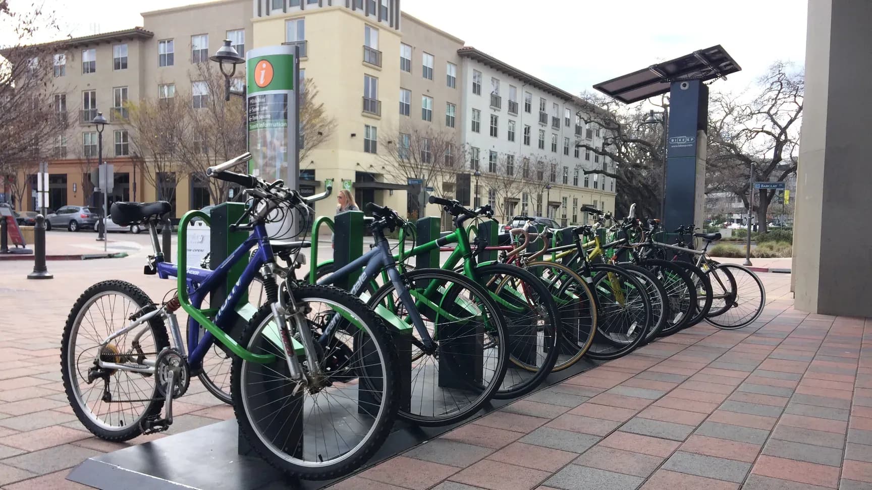 Row of bicycles locked in Bikeep smart bike parking racks beside a solar-powered station kiosk on a BART station plaza in the Bay Area.