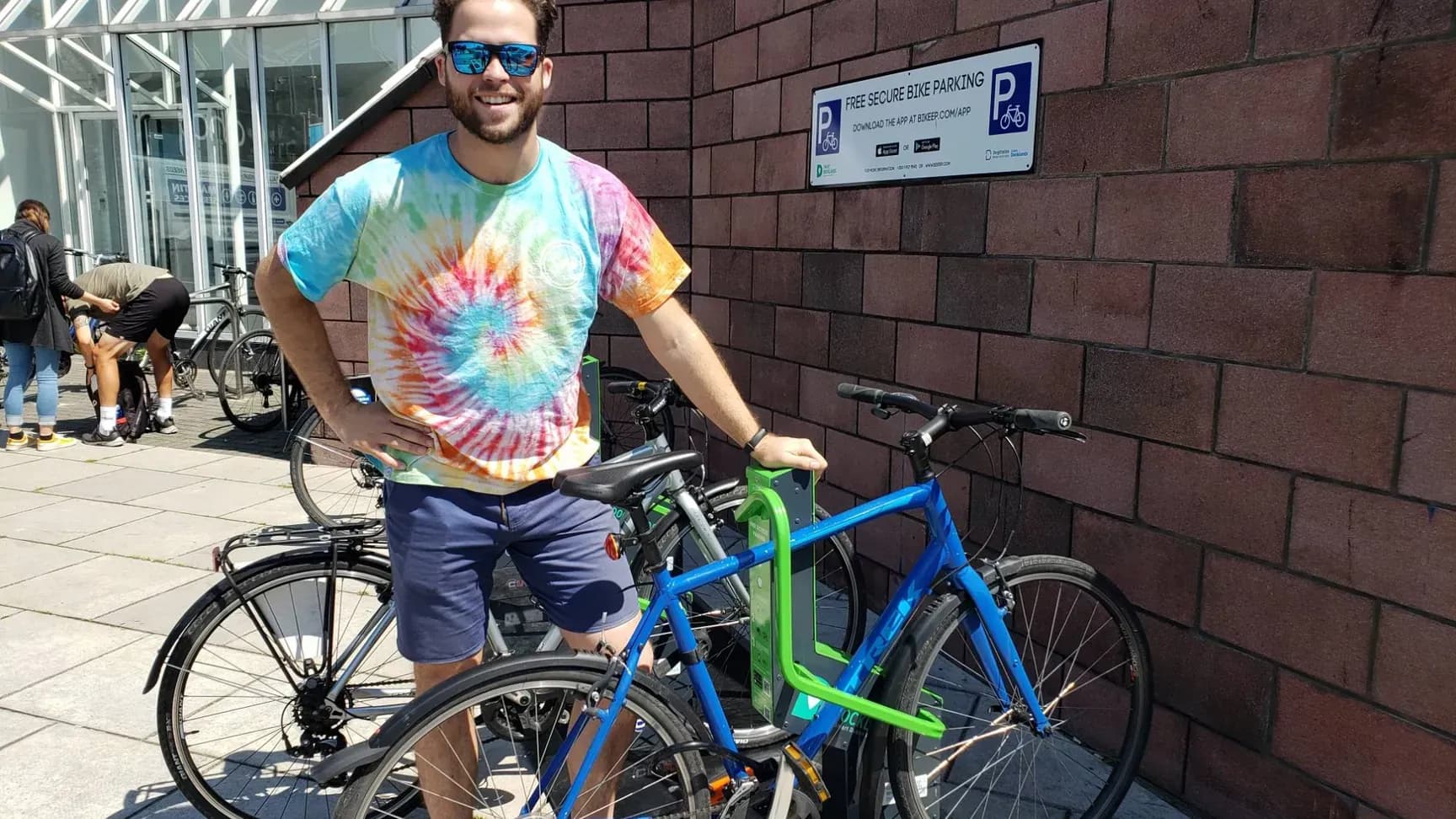 A smiling rider with a blue bike locked in a Bikeep dock beside a "Free secure bike parking" sign in Dublin's Docklands
