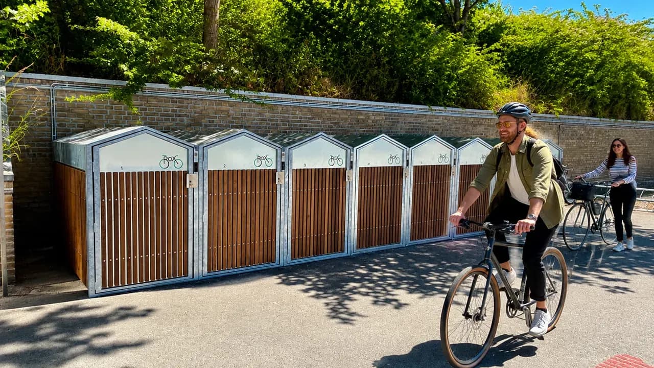 Cyclists ride past a row of smart bike lockers in Sønderborg — Nordic design in wood and galvanized steel, app-controlled with Bikeep IoT.