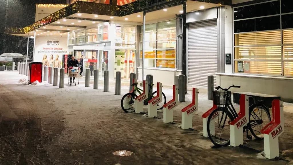 Snow-covered Bikeep bike and scooter docks outside the entrance of an ICA Maxi store in Örebro at night