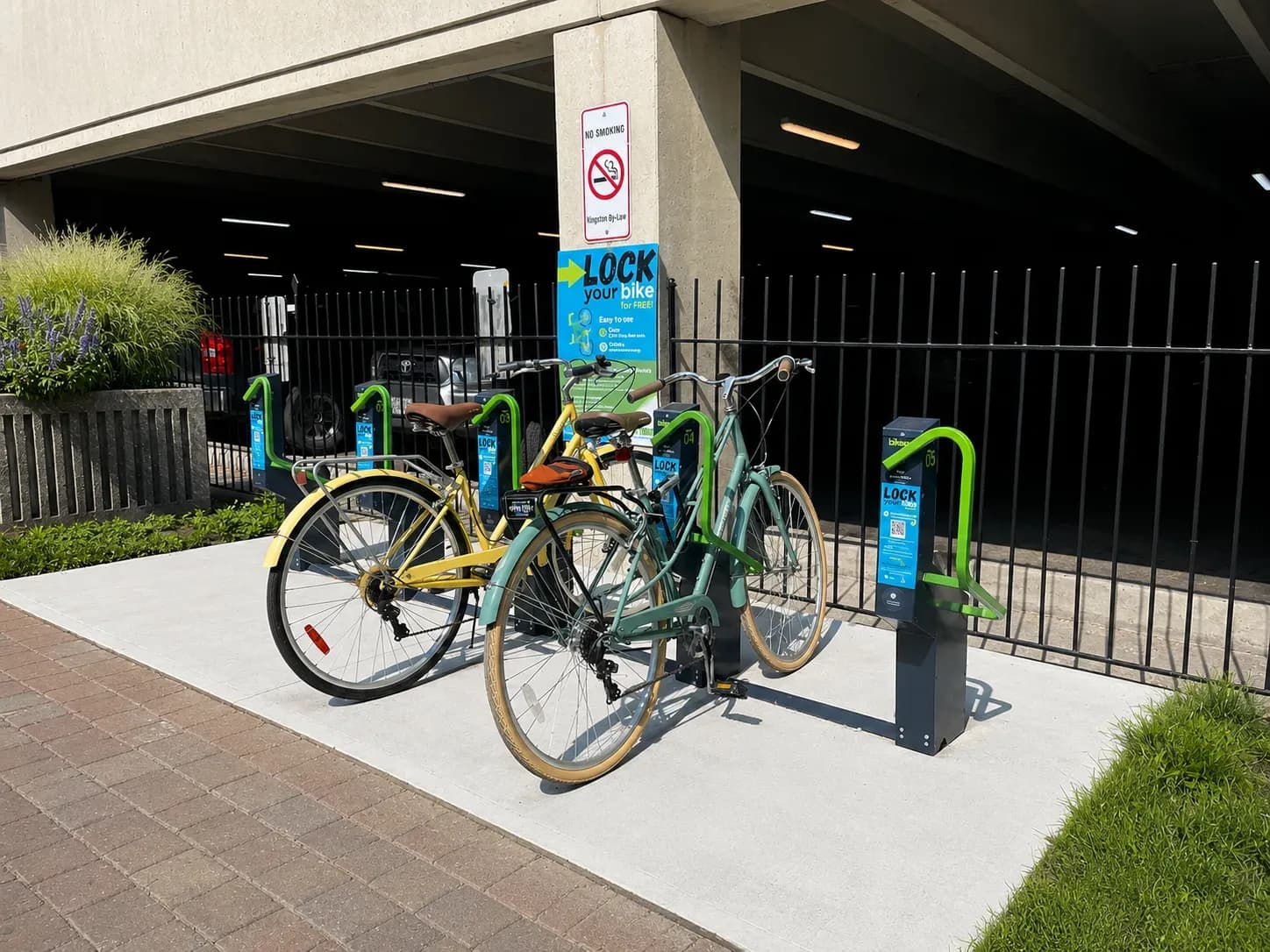 Two bicycles locked into a green Bikeep secure bike parking station in downtown Kingston, Ontario, beside a "Lock your bike" instruction sign.