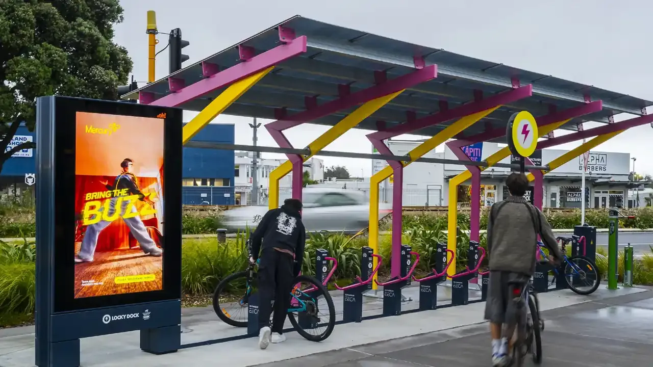 Riders docking bikes at a covered Locky Dock bike parking station beside a digital advertising screen at Paraparaumu train station, New Zealand